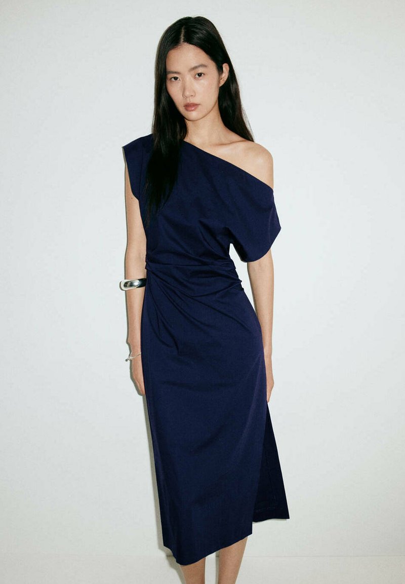 Femme aux longs cheveux noirs portant une robe midi bleu marine à une épaule avec un bracelet argenté au bras droit, debout devant un fond uni.