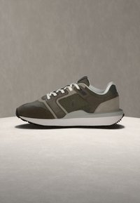 Καφέ και γκρι sneaker από δίχτυ και σουέντ με λευκά κορδόνια και χοντρή λευκή σόλα σε απλή μπεζ επιφάνεια.