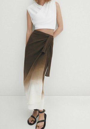Kvinde iført en hvid ærmeløs crop top, et brun-til-hvid gradient omslagsnederdel og sorte sandaler med ankelrem, står op ad en hvid væg.