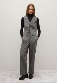 Gilet a scacchi grigi e pantaloni a gamba larga, realizzati in tessuto testurizzato. Sotto, un dolcevita nero. Scarpe nere pesanti completano l'outfit.