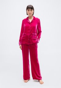 Γυναίκα στέκεται ξυπόλητη, φοράει φωτεινό ροζ βελούδινο πιτζάμα τοπ με μακριά μανίκια και κουμπιά, καθώς και ταιριαστά παντελόνια με φαρδύ πόδι, σε φωτεινό φόντο.