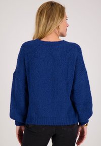 Vrouw met blond haar, gekleed in een grove, getextureerde koningsblauwe trui en zwarte jeans, met haar rug naar de camera tegen een effen achtergrond.