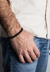 Juodų karoliukų apyrankė su lygiais, poliruotais karoliukais, dėvima ant riešo su šviesiais plaukais. Fone yra neutralus atspalvis su laisvalaikio džinsais ir marškiniais.