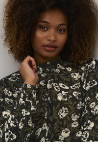 Langärmlige Bluse in Dunkelgrün mit weißem Blumenmuster, mit hohem Kragen und strukturiertem Stoff. Nahaufnahme des Ausschnitts und der Blütendetails.
