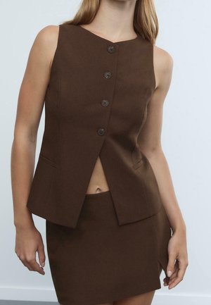 Vrouw draagt een mouwloos bruin vest met knopen en een bijpassende bruine rok met een vooraanliggende split, staand tegen een eenvoudige lichte achtergrond.