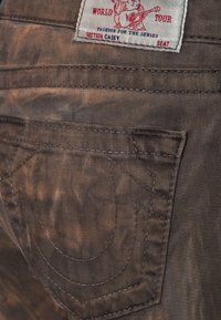 Brun denimtyg med en slitet struktur, som har en bakficka med sydda detaljer och en etikett med röd text och grafik.