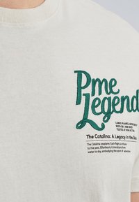 Camiseta crema con el texto bordado en verde "Pme Legend" y descripción del hidroavión Catalina. Material de algodón suave, cuello redondo clásico.
