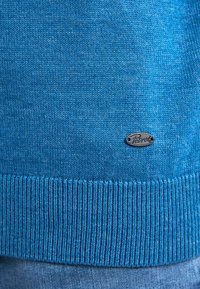 Blaues Strickpullover mit geripptem Saum und einem kleinen silbernen Logopatch auf der Vorderseite, aus einem weichen, strukturierten Material gefertigt.