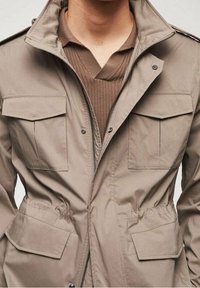 Veste légère beige dotée d'une fermeture éclair, de deux poches poitrine et de deux poches latérales, avec des détails élastiques à la taille.