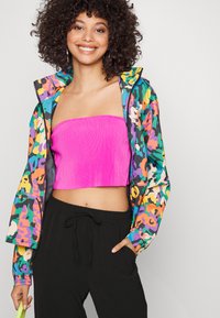 Giacca a hood colorata con un vivace motivo astratto, abbinata a un crop top rosa a coste e pantaloni neri morbidi, che mostra uno stile casual.