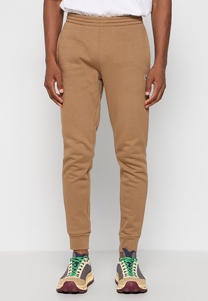 Person iført brune, tætsiddende joggebukser og flerfarvede sneakers med grønne snørebånd, stående mod en ensfarvet lys baggrund.