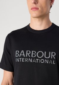 T-shirt de algodão preto com um design de logotipo texturizado em branco e prateado, com decote redondo e mangas curtas. Estilo minimalista.