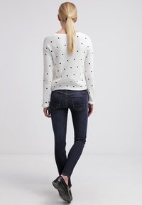 Suéter blanco de canalé con lunares negros, combinado con jeans ajustados de color azul oscuro. El look incluye zapatos negros, visto desde la parte de atrás.