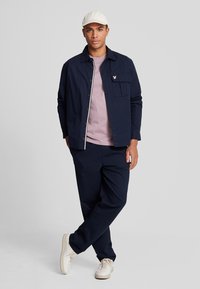 Giacca di cotone blu navy con cerniere bianche e tasca sul petto, abbinata a una maglietta rosa chiaro e pantaloni blu navy, completata da sneaker bianche.