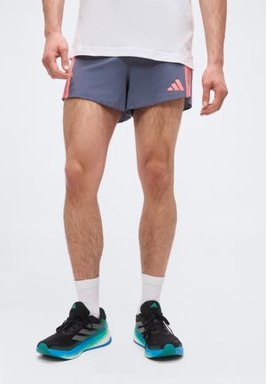 Muž nosící šedé šortky Adidas s růžovými pruhy a logem, bílé ponožky a černé běžecké boty s tyrkysovými a modrými detaily.
