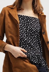 Brauner Wildleder-Trenchcoat mit Reverskragen, kombiniert mit einem schwarzen Blumenoberteil mit Peplum-Saum und dünnen Trägern.