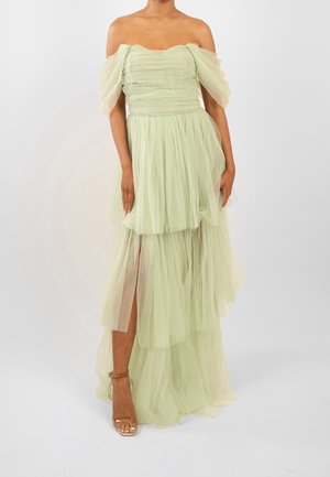 Kobieta w bladziezielonej, warstwowej sukni z tiulu z odkrytymi ramionami i wysokim rozcięciem, w złotych sandałach na cienkich paskach, pozująca na jednolitym tle.