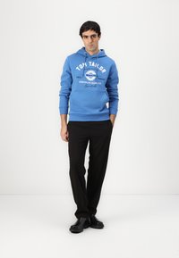 Blaues Sweatshirt mit weißen Drucktexten und Logo, kombiniert mit schwarzen Hosen und schwarzen Stiefeln. Glatte Textur, lässige Passform und Kapuze mit Kordelzug.