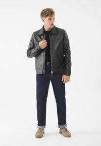 Zwarte leren jas met een kraag, voorrits en twee zijzakken, gecombineerd met donkerblauwe jeans en beige schoenen. Gladde textuur.