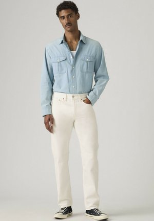 Uomo con camicia celeste chiara abbottonata, pantaloni bianchi e scarpe da ginnastica nere, in piedi con una mano in tasca su sfondo semplice.