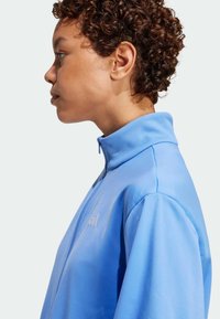 Svetlo modra športna jakna z visokim ovratnikom in polovičnim zadrgo, ki se ponaša z gladko teksturo ter diskretnim logotipom na prsih.