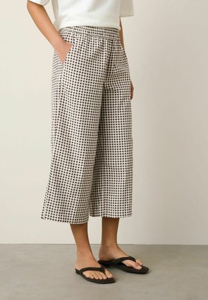 Personne portant un pantalon large court à carreaux noir et blanc avec taille élastique et tongs noires, debout avec une main dans la poche.
