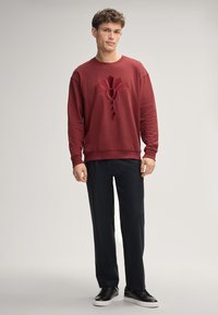 Felpa bordeaux con design a cavallo, vestibilità comoda, polsini e orlo a coste, abbinata a pantaloni scuri e sneaker nere eleganti.
