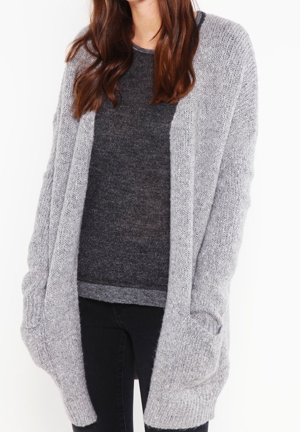 Personne portant un cardigan en tricot gris clair, ample, par-dessus un pull gris foncé et un pantalon noir, sur un fond blanc.
