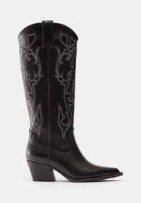 Botte de cowboy haute en cuir noir avec des coutures blanches décoratives et un talon empilé incliné.