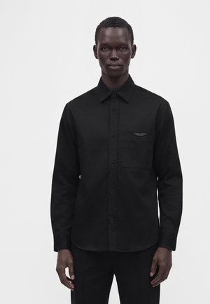 Homme portant une chemise noire à boutons avec une poche poitrine et un petit emblème ailé, debout devant un fond blanc uni.
