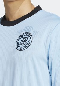 Ljusblått idrottströja med ribbad textur, svart rund halsringning och broderad cirkulär logotyp med text och designdetaljer.