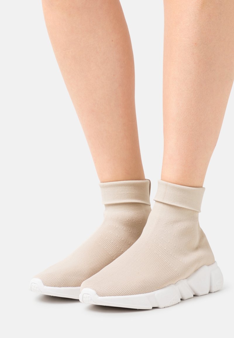 Pieds portant des baskets style chaussette beiges avec des semelles épaisses blanches et volumineuses sur fond blanc.