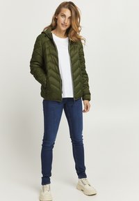 Podložena olivno zeleni jakna s chevron vzorcem, z zadrgo spredaj in kapuco, oblečena čez enostavno belo srajco in modre jeans hlače.