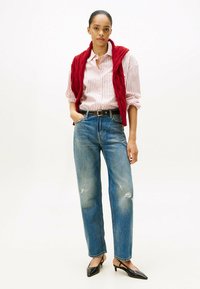 Chaleco acolchado rojo sobre una camisa de botones a rayas, combinado con jeans de mezclilla azules desgastados y tacones negros tipo slingback. Un look sencillo y casual.