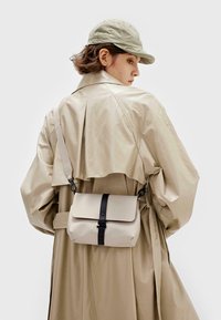 Beige Umhängetasche mit Klappenverschluss und einem schwarzen Riemen, getragen über einem cremefarbenen Trenchcoat mit strukturiertem Kragen und fließenden Ärmeln.