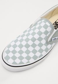 Baskets slip-on avec motif damier gris clair et blanc, semelle en caoutchouc blanche et étiquette logo Vans sur le côté.