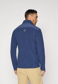 Giacca in pile navy con superficie testurizzata, colletto alto e pannelli a contrasto sulle spalle. Presenta un logo sulla parte superiore della schiena.