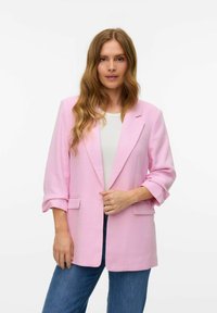 Blazer rosa con una tela texturizada, collar tipo solapa, mangas enrolladas y bolsillos frontales, combinado con una camisa blanca y jeans azules.