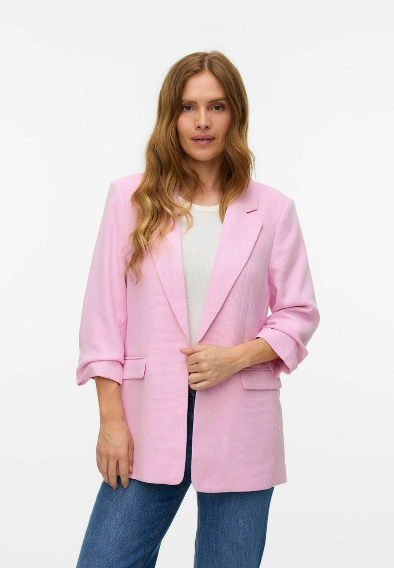 Blazer rosa con una tela texturizada, collar tipo solapa, mangas enrolladas y bolsillos frontales, combinado con una camisa blanca y jeans azules.