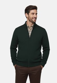 Sweater oscuro verde con cremallera y cuello alto, de tela texturizada, con puños acanalados, llevado sobre una camisa de cuadros, combinado con pantalones marrones.