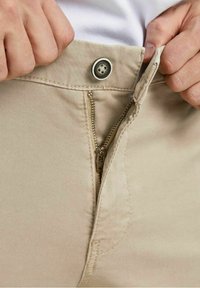 I pantaloni beige presentano una chiusura con zip, fissata con un bottone rotondo in vita. Il tessuto è lucido, con una finitura pulita e cuciture visibili.