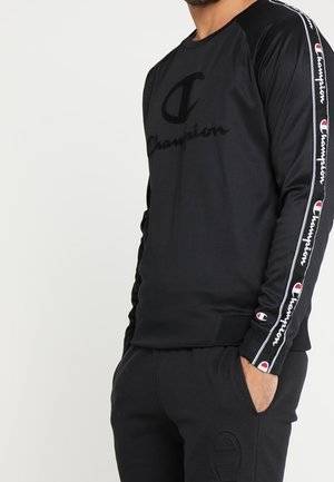 Mann trägt schwarzen Champion-Sweatshirt mit Logo auf der Brust und markanten Streifen entlang der Ärmel, Hände in den Taschen einer schwarzen Jogginghose.