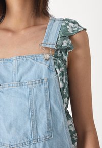 Ανοιχτό μπλε τζιν σαλοπέτα με μεγάλη μπροστινή τσέπη, ασημένια αξεσουάρ και ρυθμιζόμενους ιμάντες, φορεμένη πάνω από ένα πράσινο φλοράλ τοπ με φουσκωτά μανίκια.