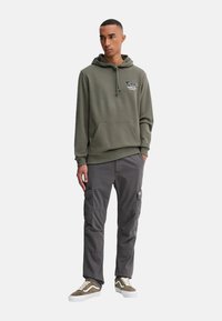 Sudadera verde oliva con bolsillo frontal y cordón, combinada con pantalones cargo grises con bolsillos laterales y zapatillas marrones y blancas.