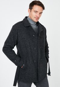 Cappotto in lana di un grigio scuro, con una texture, doppio petto, due bottoni e tasche laterali. Presenta un colletto e passanti per cintura regolabili.