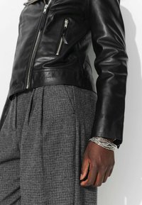 Veste en cuir noir avec des détails de fermeture éclair argentés, associée à un pantalon plissé à carreaux gris. Un bracelet en argent est porté au poignet.