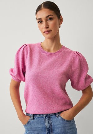 Kvinde med bagstrøget hår iført en lyserød sweater med pufærmer og blå jeans, stående med hænderne i lommerne mod en ensfarvet baggrund.