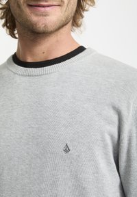 Jasnoszary sweter z miękkiej tkaniny. Ma czarną ściągaczkę przy dekolcie i małe haftowane logo na piersi. Klasyczny krój.