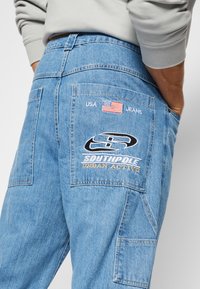 Hele hele sinise denimpüksid, millel on tagasi tasku, millel on broderitud logo, USA tekst ja Ameerika lipu plaastr. Klassikaline lõige, küljetaskud.