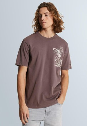 Jeune homme aux cheveux bruns ondulés portant un T-shirt marron avec des motifs floraux blancs et le texte « Cast Iron », pantalon gris clair, debout devant un fond uni.
